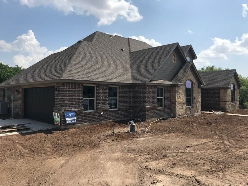 New brick house under construction with a gray roof on a brown dirt lot under a blue sky.