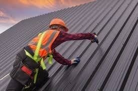Roofer in safety gear installing metal roofing on a slanted roof at sunset.