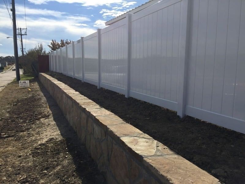 A white vinyl fence on top of a low stone retaining wall along a roadside.