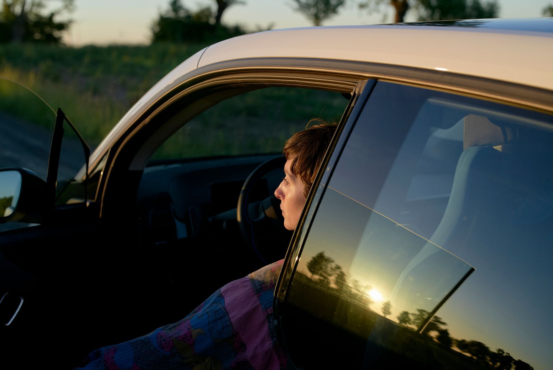 A woman is sitting in a car looking out the window at the sunset.