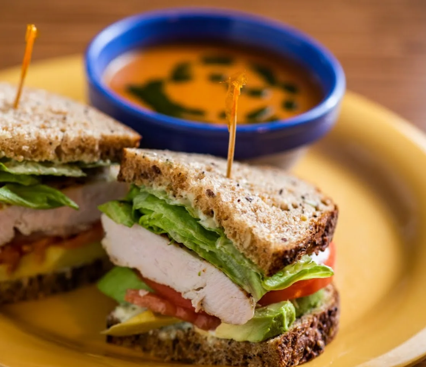 Sandwich halves with chicken, tomato, lettuce on wheat bread; served with tomato soup in a blue bowl, on a yellow plate.