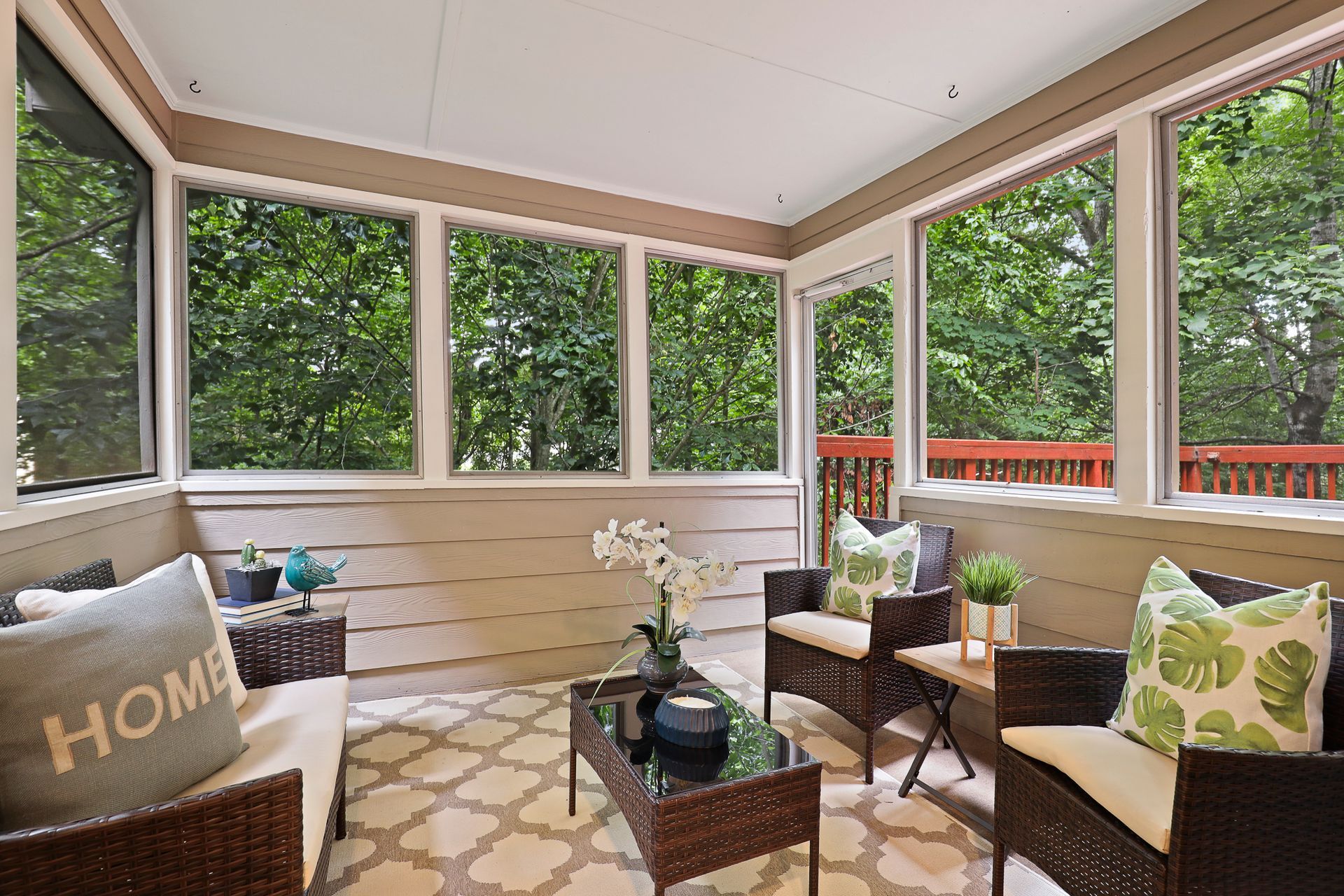 A sunroom with wicker chairs, a small table, a patterned rug, and large windows overlooking green trees.
