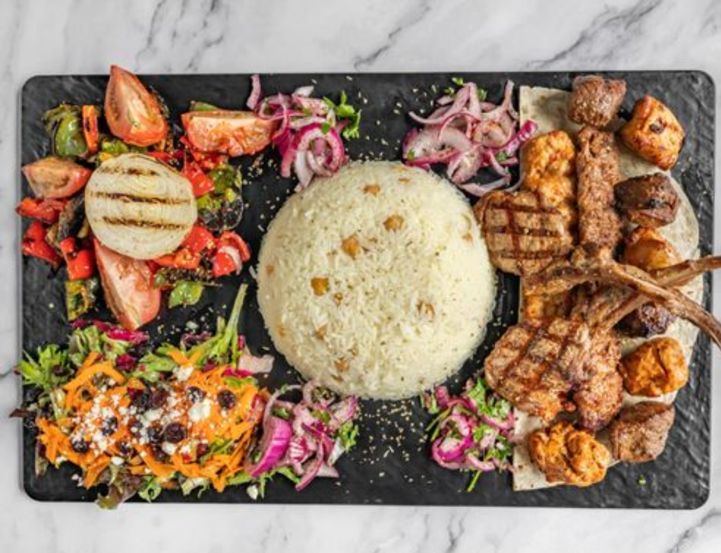 A platter of grilled meats, rice, and vegetables, including tomatoes, onions, and carrots, on a black slate.
