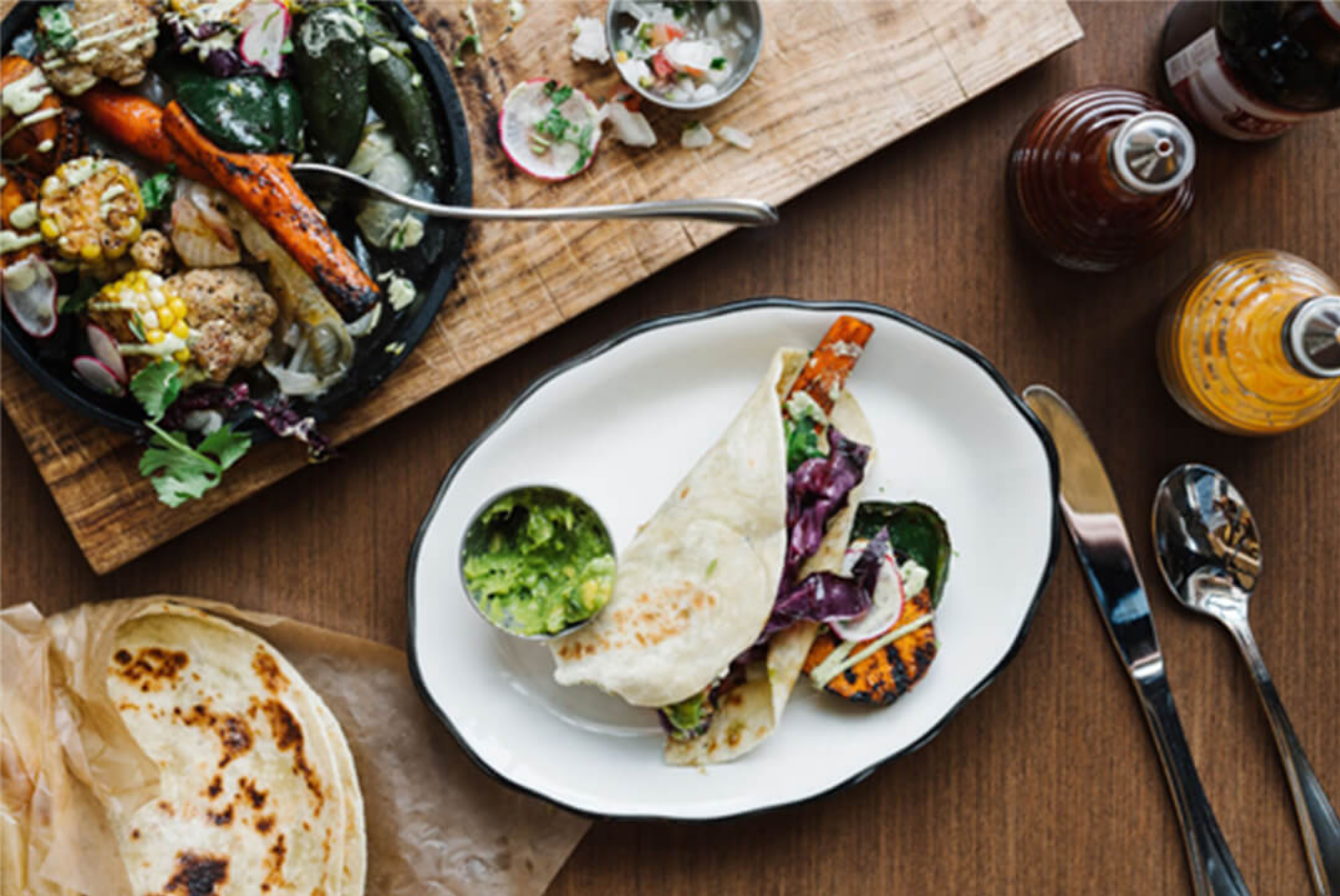 Taco with grilled vegetables, guacamole, and tortillas on a wooden table with sauces and utensils.