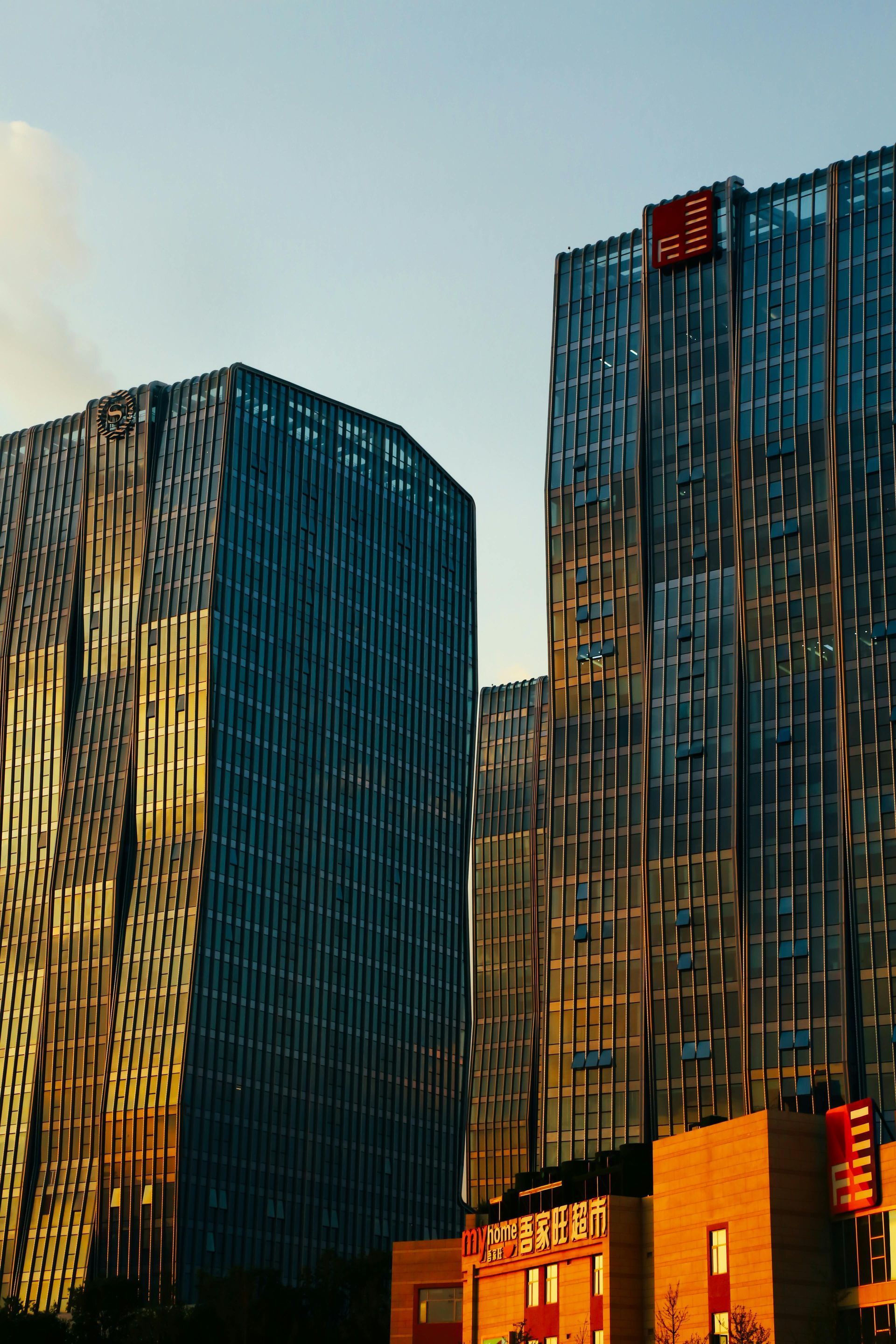 Two modern skyscrapers with glass facades at sunset, illuminated with warm, golden light against a pale blue sky.