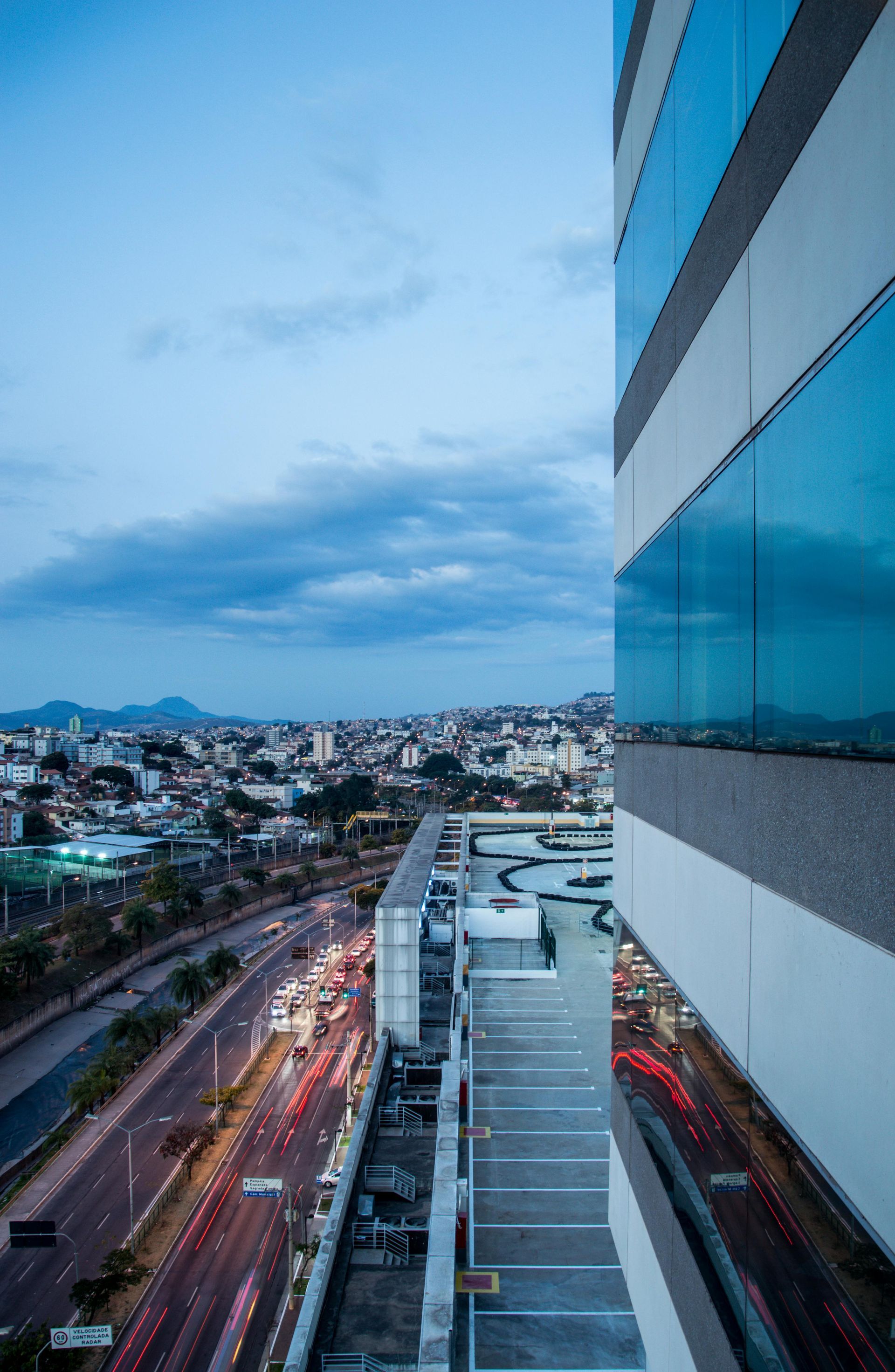 High-angle view of a city street at twilight, with light trails from traffic alongside a modern glass-clad skyscraper.