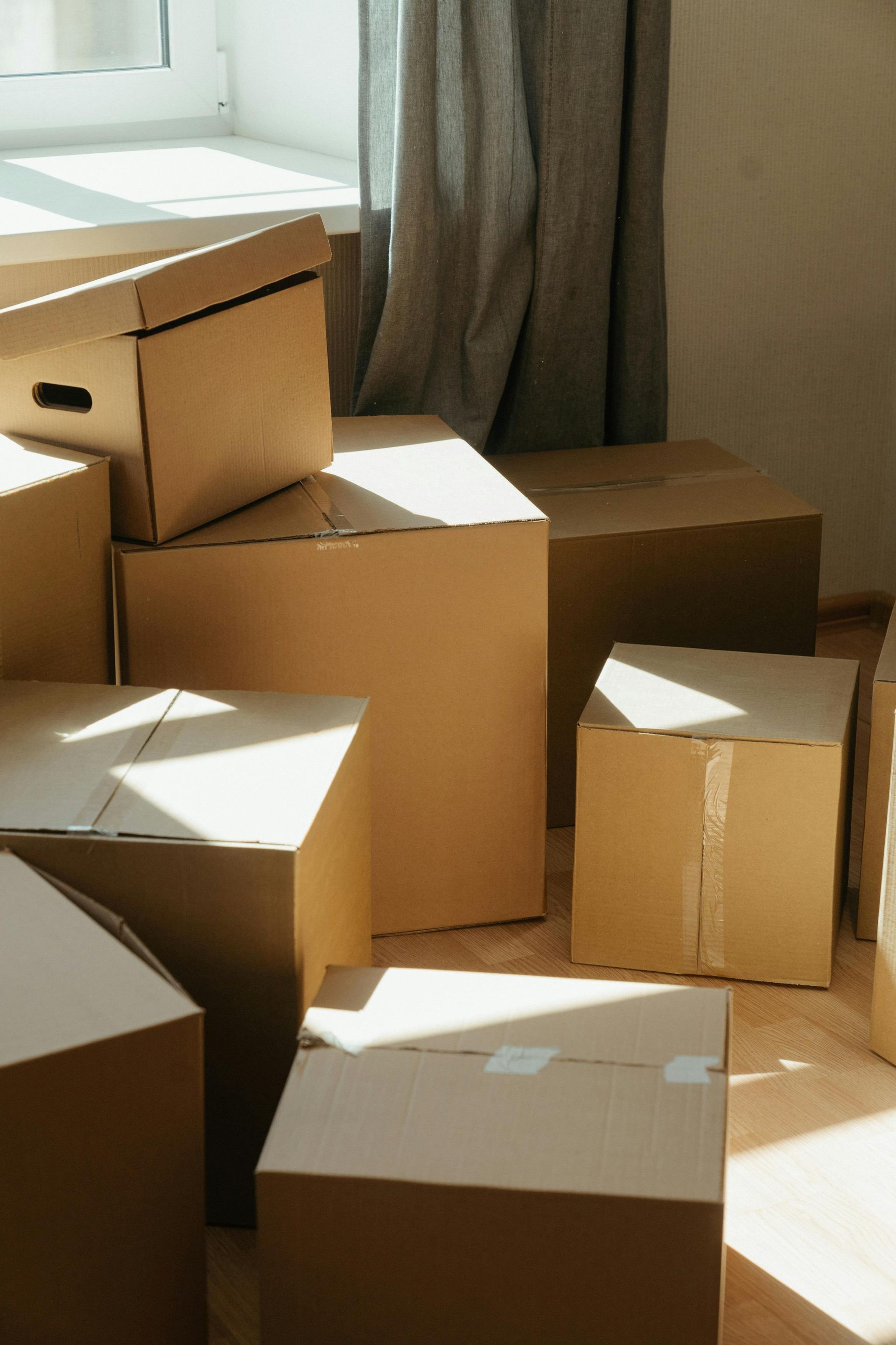 A stack of brown cardboard moving boxes sits on a wooden floor in a brightly lit room near a window and curtains.