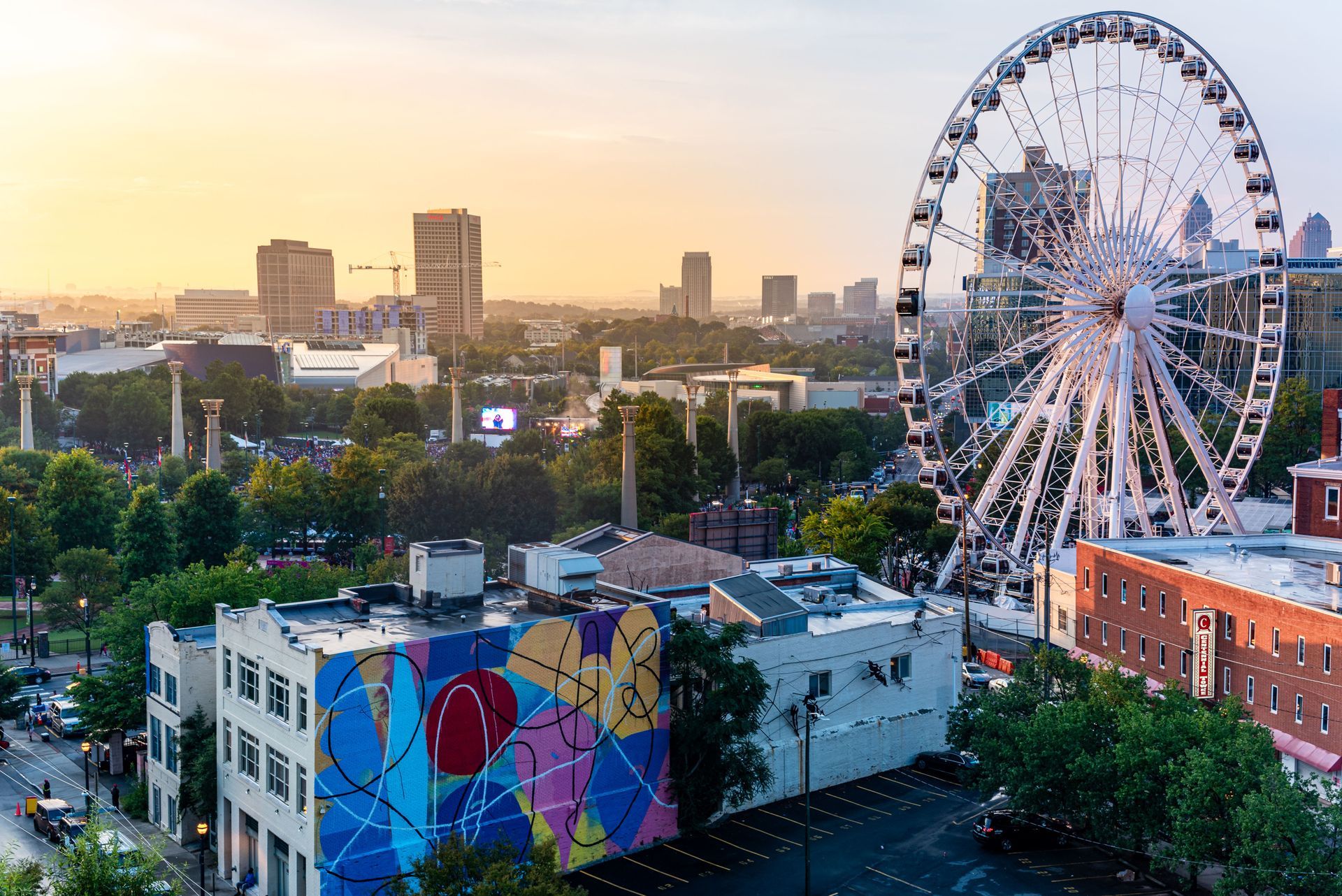 Ferris wheel above city buildings at sunset, with a colorful mural-covered rooftop in the foreground