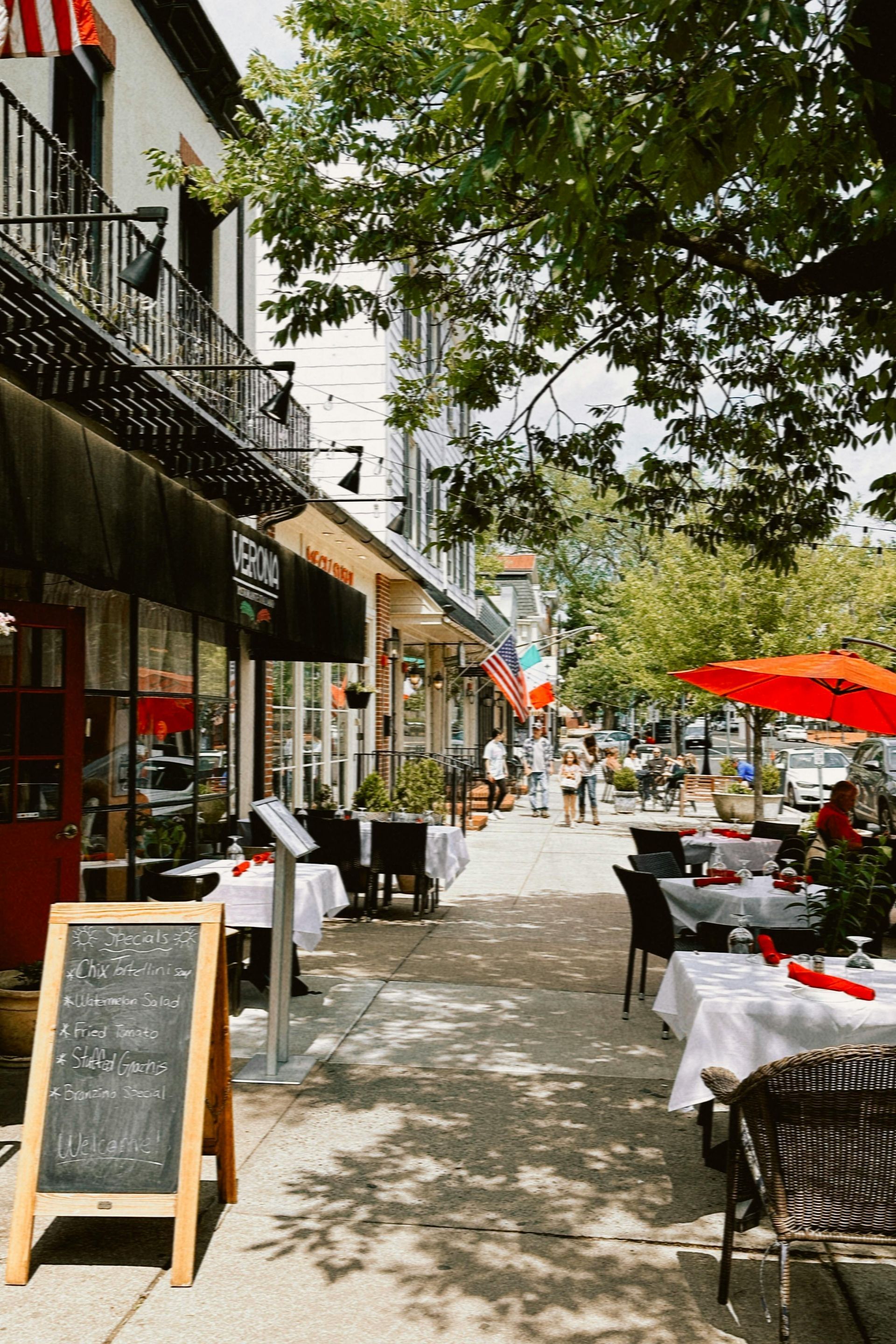 Outdoor dining along a sunny, tree-lined street with sidewalk tables, umbrellas, and a chalkboard menu.