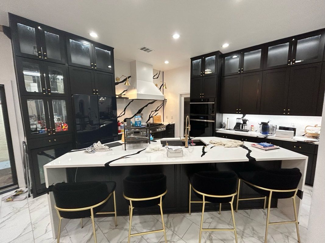 A kitchen with black cabinets , white counter tops , and gold stools.
