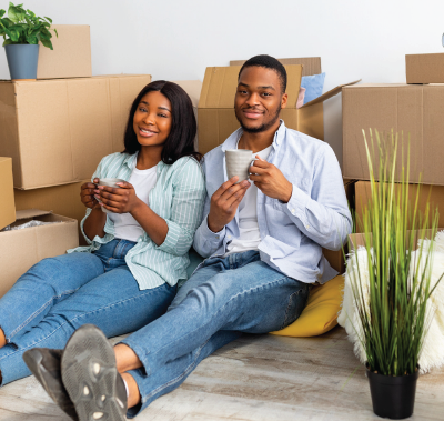 Couple sitting among moving boxes, drinking from mugs, smiling.