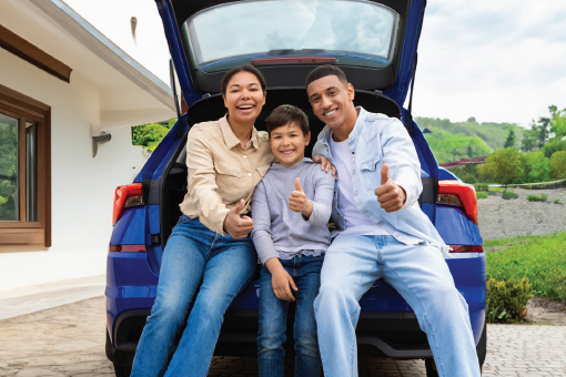 Three people sitting in an open car trunk outdoors, smiling and giving thumbs up.