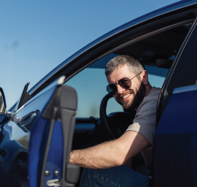 Man in sunglasses smiles from a blue car, door open, clear sky.