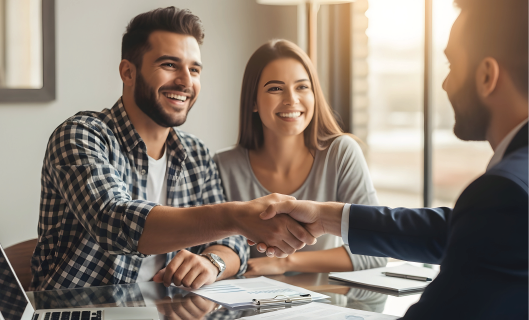 A professional interaction where a person in a plaid shirt shakes hands with someone in a suit while a companion smiles.