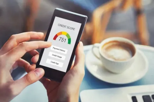 Hands holding a smartphone showing a credit score of 751 on a screen, sitting near a coffee cup and laptop.