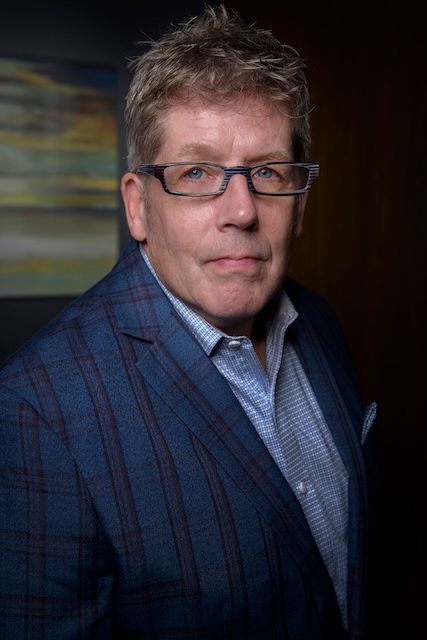 Man with glasses in blue plaid blazer, light blue shirt, looking forward in a room.