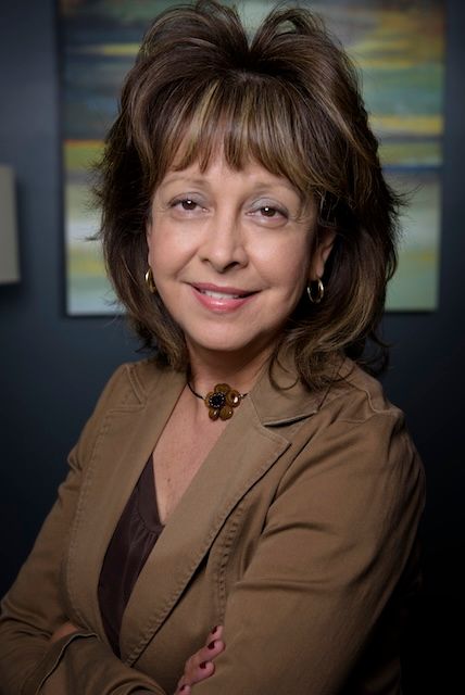 Woman in a brown jacket smiles, arms crossed. Dark hair, background art, dark wall.
