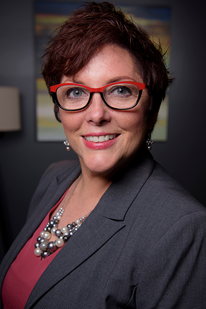 Woman in red-rimmed glasses, blazer, and pearl necklace smiles, posing indoors near a painting.