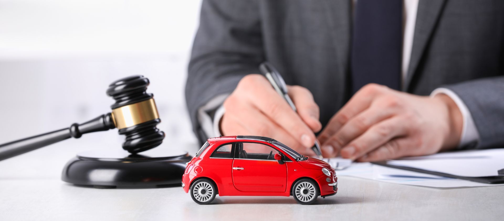 A person in a suit signing paperwork with a red toy car and gavel on the table.