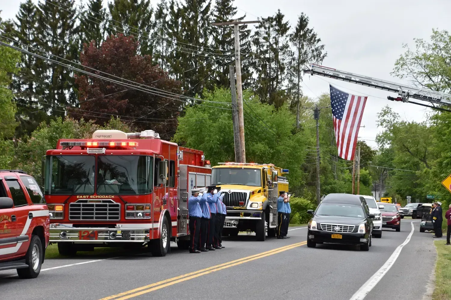 Fire trucks and vehicles in a procession; firefighters saluting next to a hearse. American flag draped.