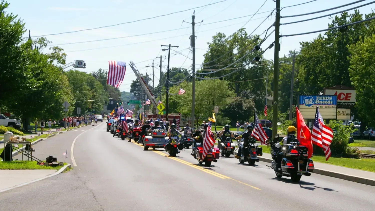 Motorcycle parade with American flags on a sunny road, near an Ace Hardware store.