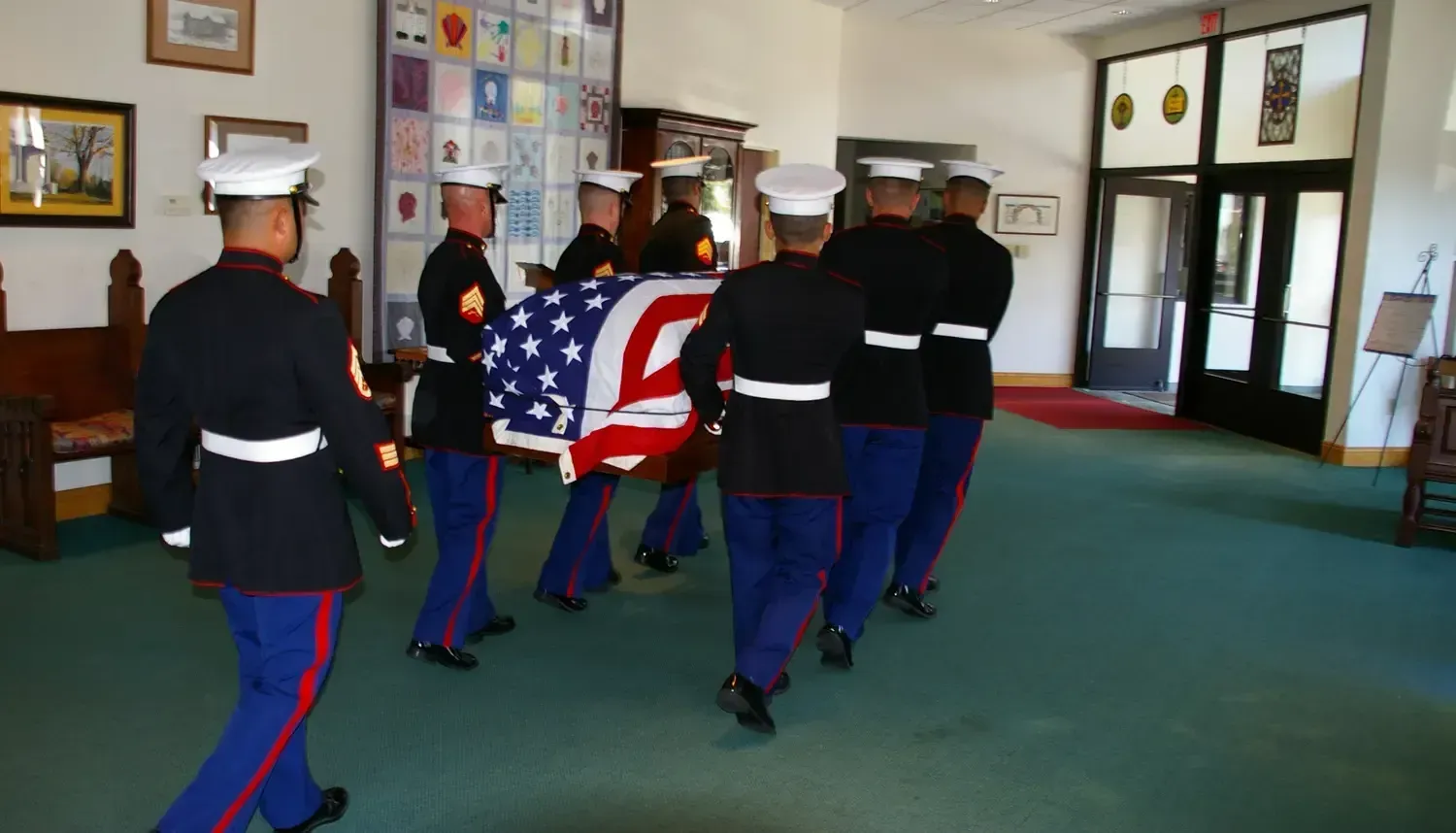 Marines carry a flag-draped casket indoors, likely a funeral service, wearing uniforms.