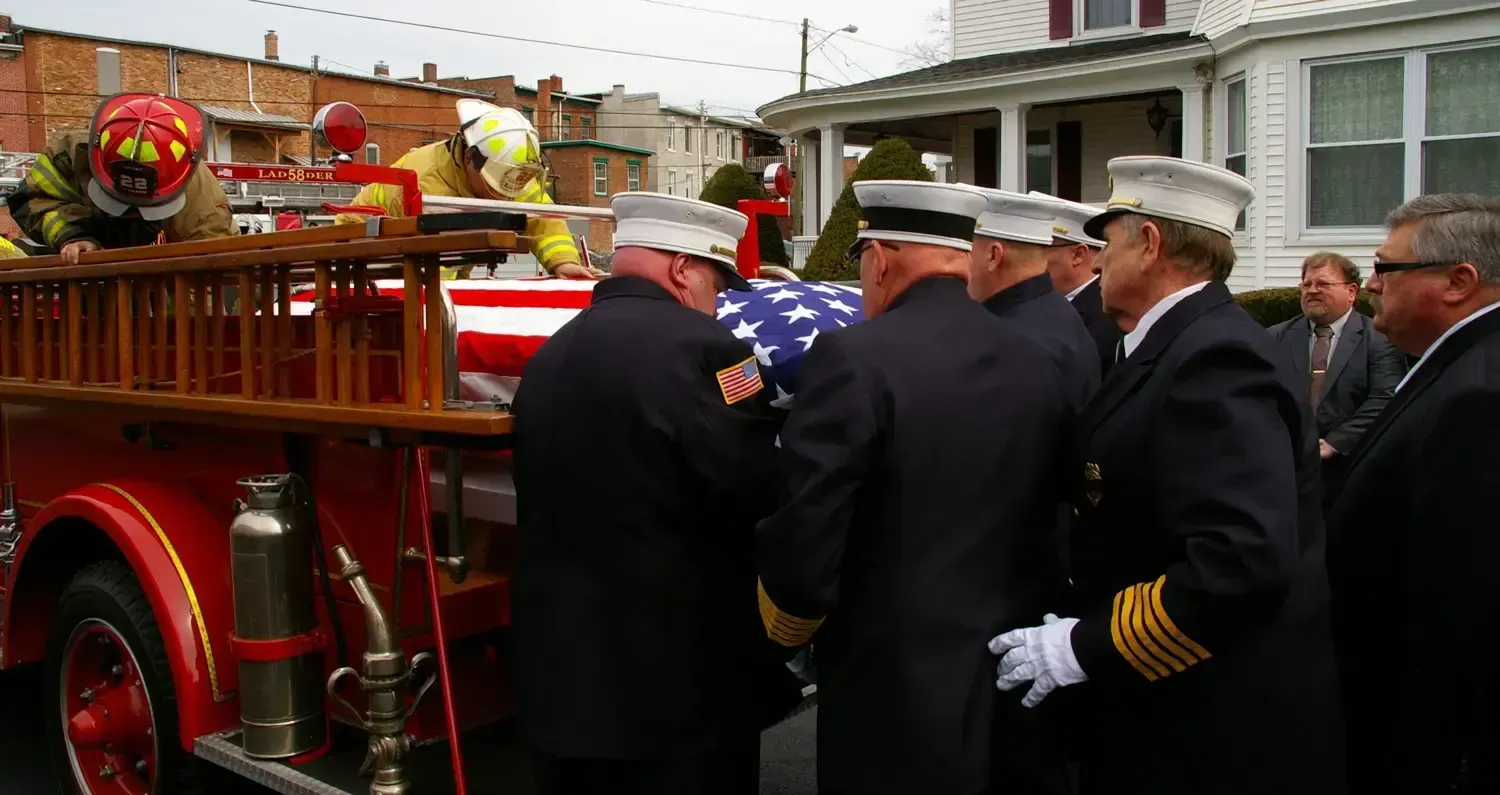 Firefighters carrying a flag-draped casket on a vintage fire truck. Other men in uniforms stand nearby.