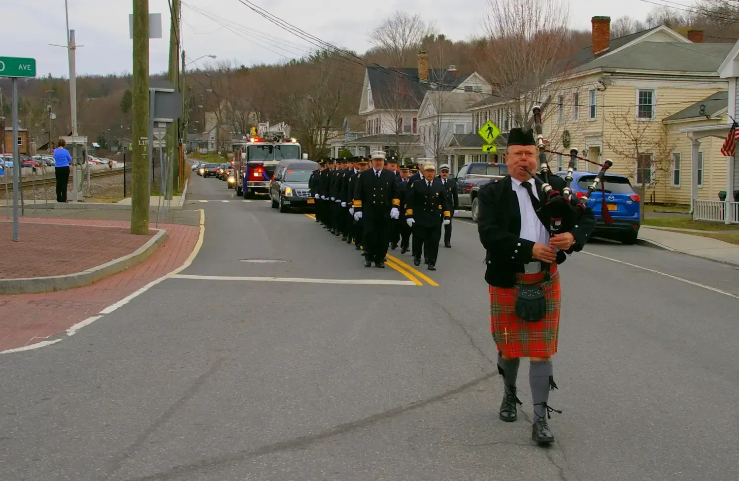Bagpiper leading a procession of uniformed firefighters, vehicles, and onlookers down a street.