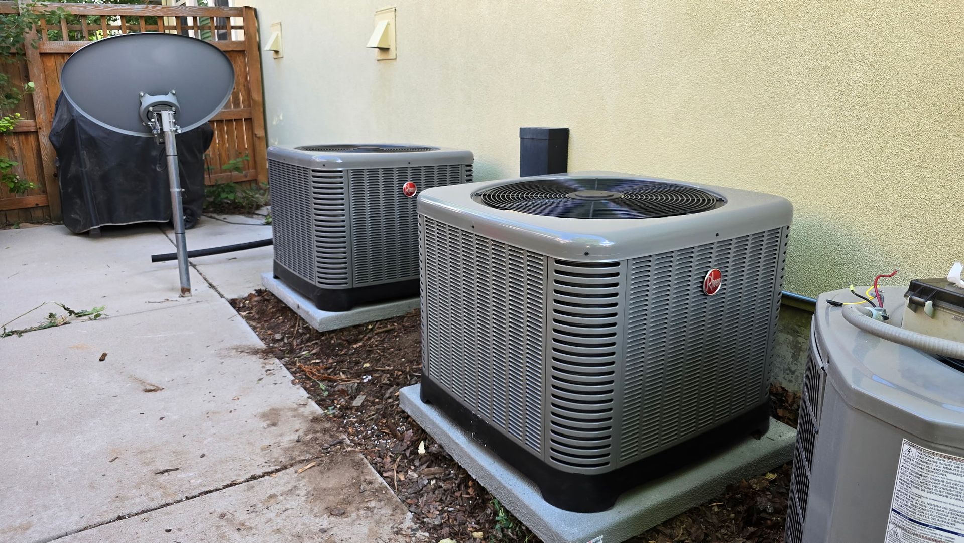Two air conditioners are sitting outside of a house next to a satellite dish.
