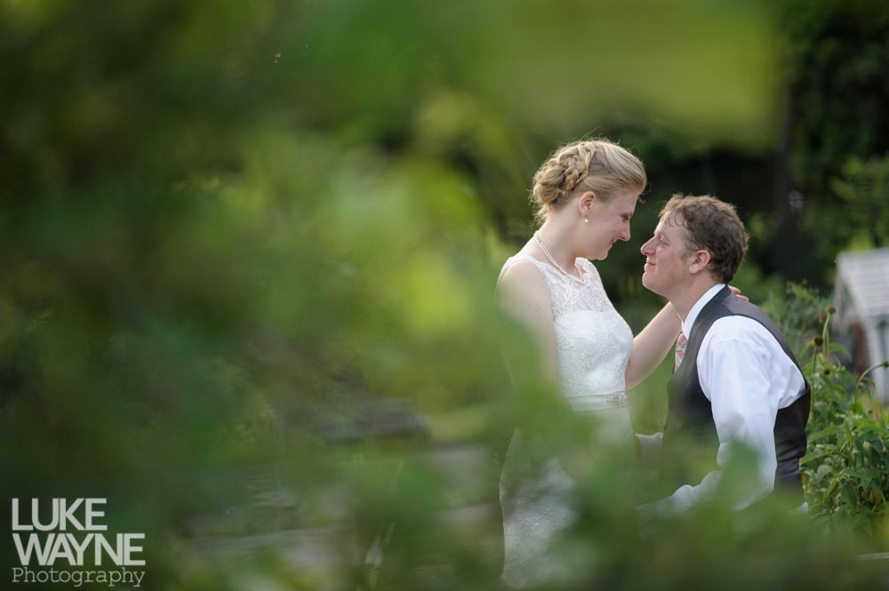 Bride and groom face each other, touching faces, through green foliage. They smile. Outdoor setting.