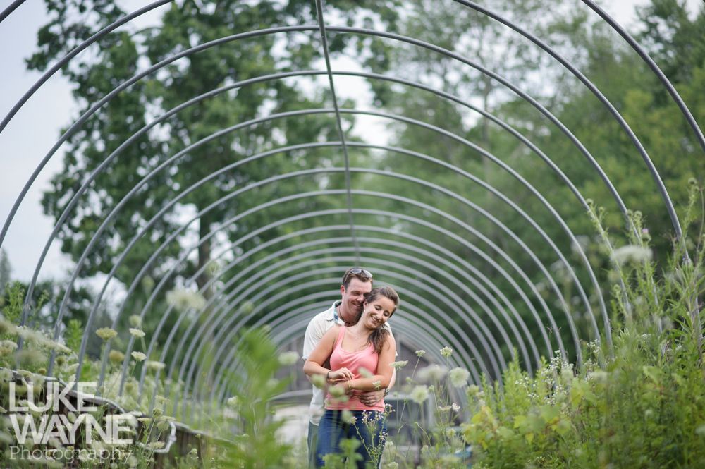 Couple embraces under a metal archway in a garden; woman smiles, man's arms around her waist.