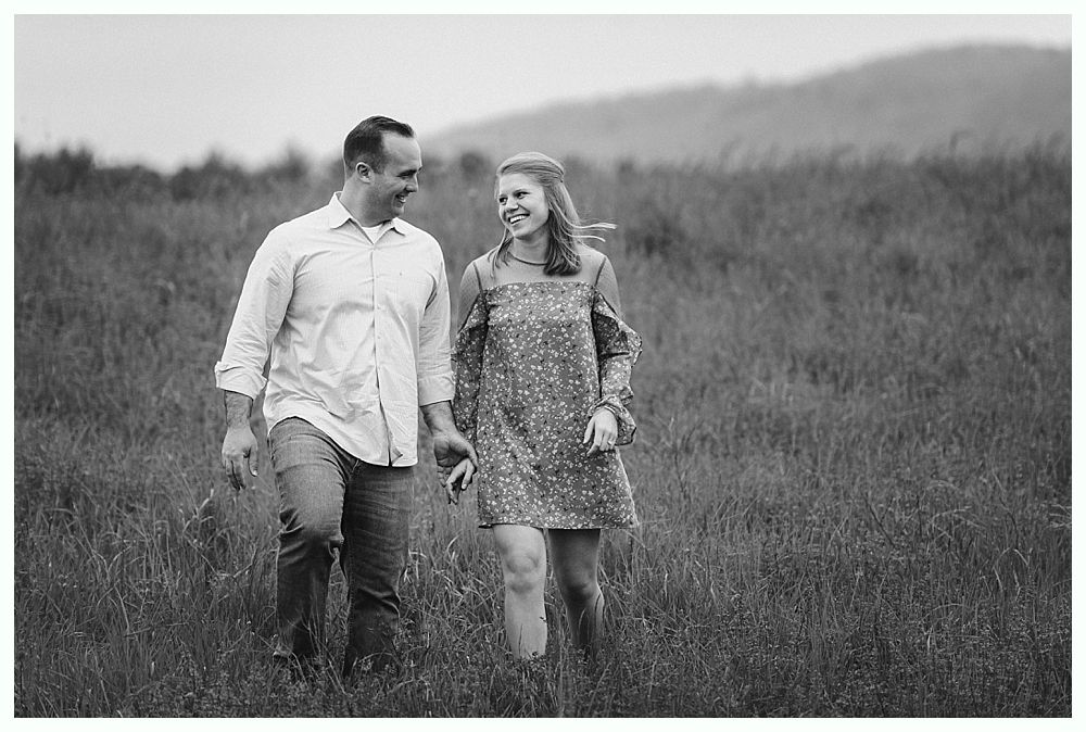 Couple holding hands, smiling at each other while walking through tall grass in a field.
