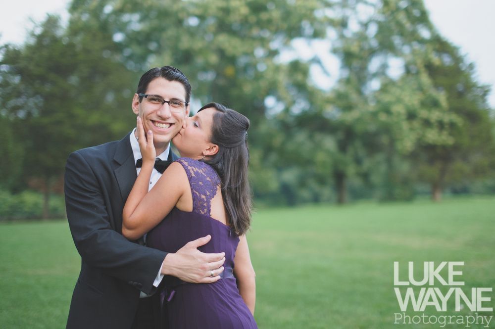 Woman kisses man's cheek outdoors. She wears a purple dress, and he wears a tuxedo. They smile.