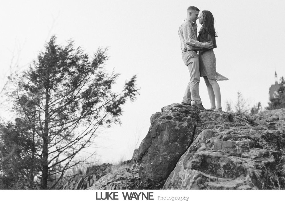 Couple embraces on a rocky outcrop, facing each other. Black and white photograph, cloudy sky, tree on the left.