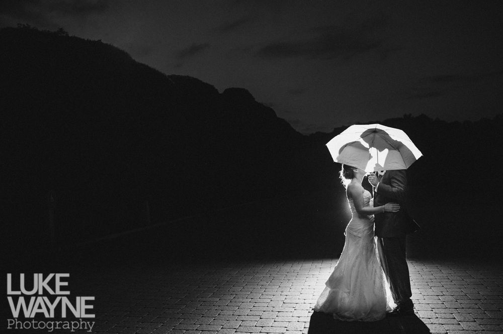 Bride and groom under umbrella, backlit by light. Mountain backdrop in dark, black and white photo.