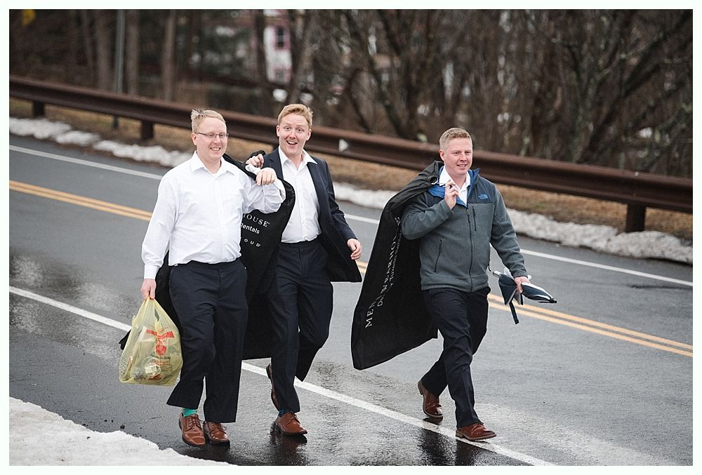 Groom smiles at camera, surrounded by groomsmen in navy suits and burgundy ties, outdoors.