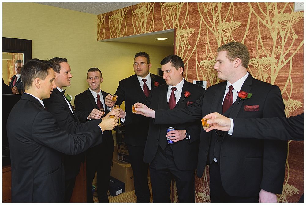 Groom smiles at camera, surrounded by groomsmen in navy suits and burgundy ties, outdoors.