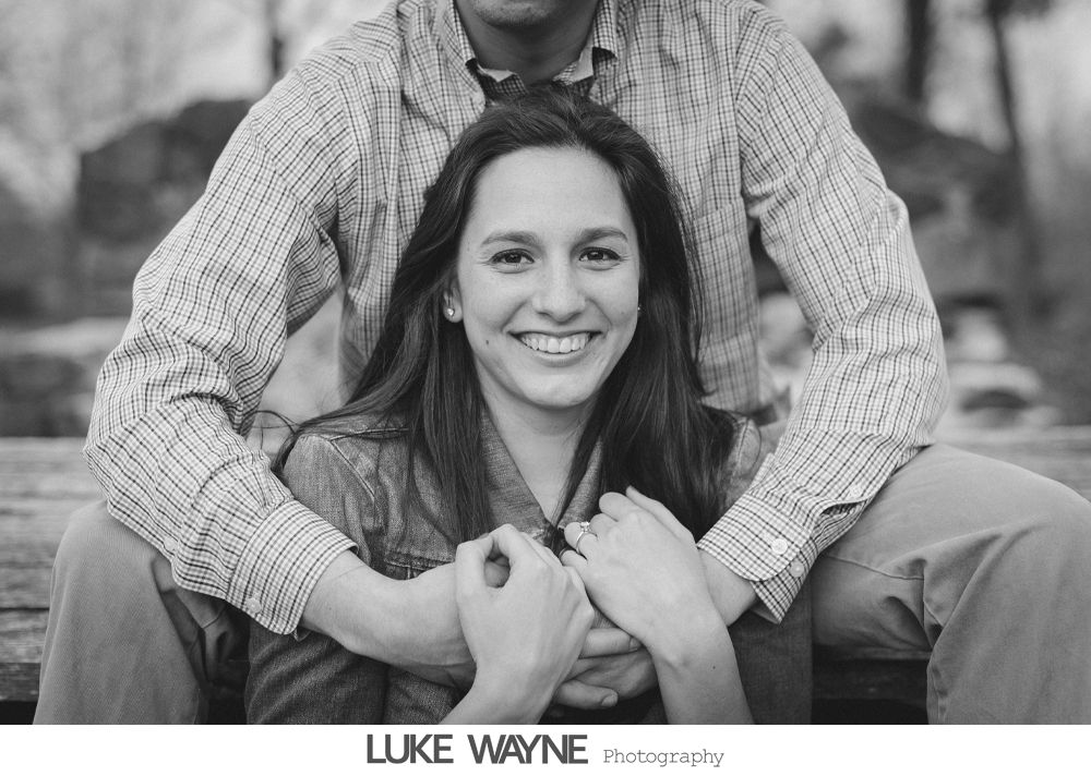 Couple embracing, woman smiling, man's arms around her. Black and white photo, outdoor setting.