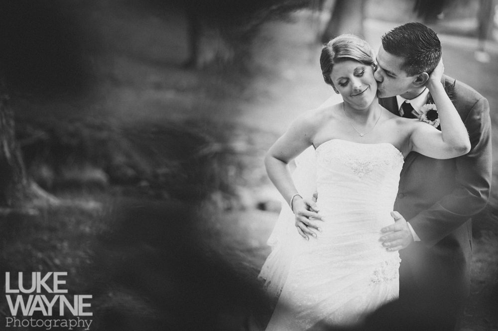 Bride and groom embrace outdoors, groom kisses bride's cheek. Black and white photo with blurred forest background.