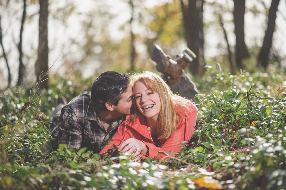 Bride and groom embrace outdoors, groom kisses bride's cheek. Black and white photo with blurred forest background.
