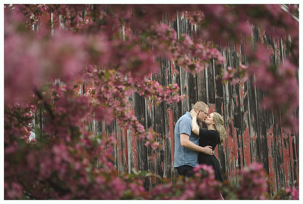 Bride and groom embrace outdoors, groom kisses bride's cheek. Black and white photo with blurred forest background.