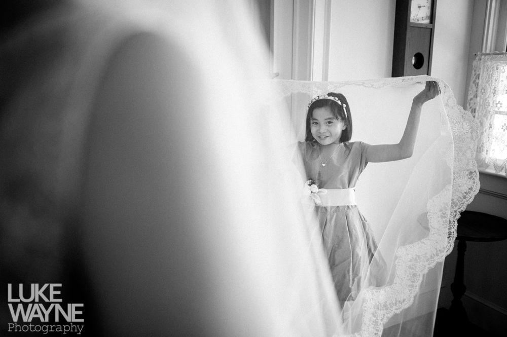 Girl holding a wedding veil, smiling. Bride in the foreground is out of focus. Interior, black and white.