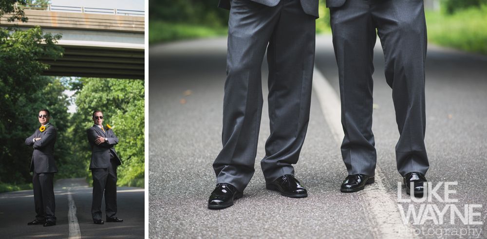 Two men in suits stand on a road. One has arms crossed, a yellow tie. The other is beside him, also in a suit, with dark sunglasses.