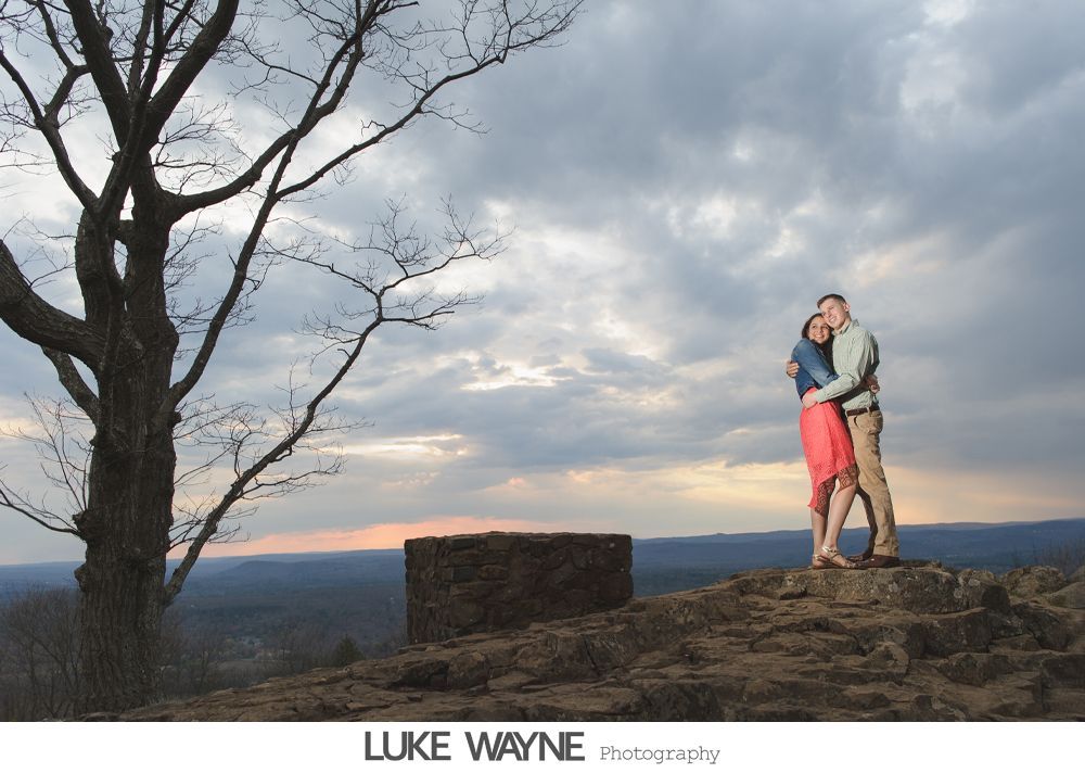 Couple embraces on a rocky hilltop, silhouetted tree, sunset, dramatic sky.