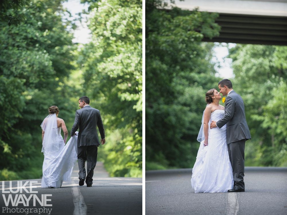Bride and groom in wedding attire walking and kissing on a tree-lined road.