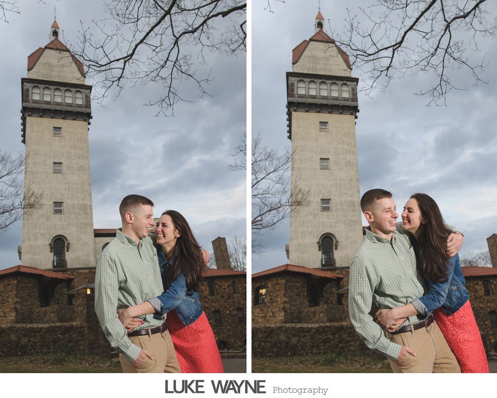 Couple embracing, smiling in front of a tall stone tower with cloudy sky background.