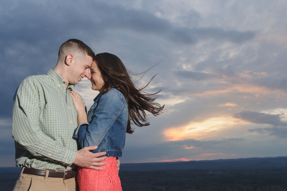 Couple embracing at sunset; man in green shirt, woman in denim jacket and red dress, with blowing hair.