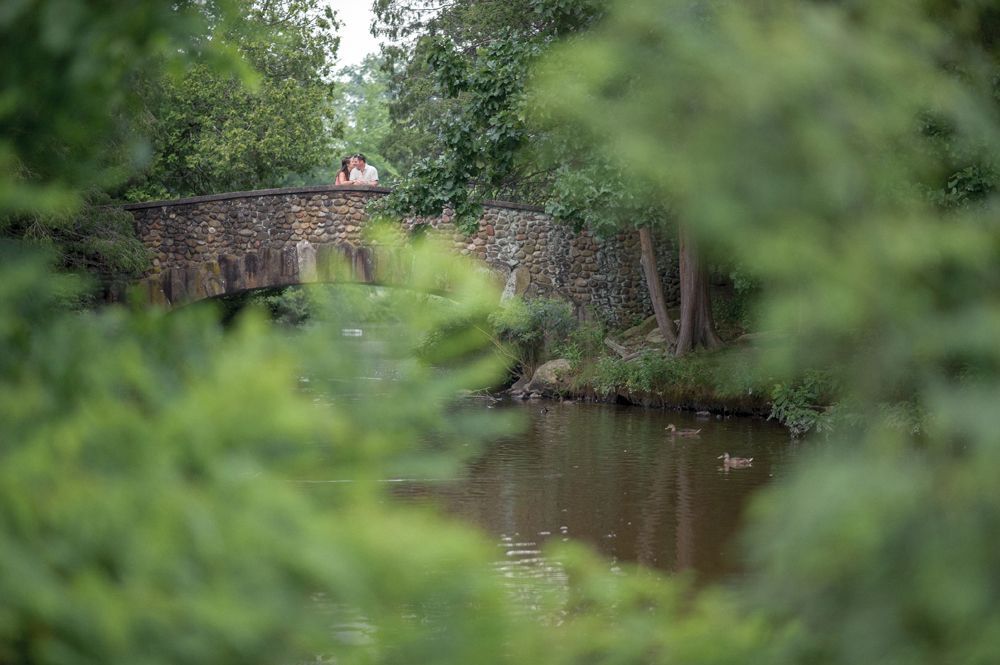 Stone bridge over a calm river, partially obscured by green foliage; two people stand on the bridge.