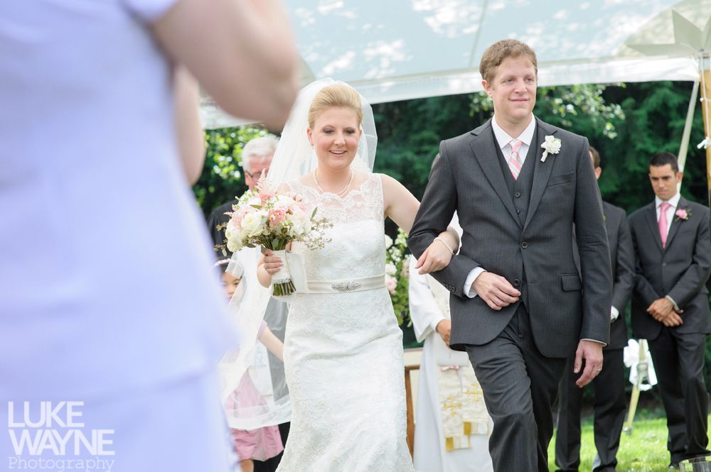 Bride and groom exiting wedding ceremony arm in arm, smiling. Outdoor setting, other attendees present.