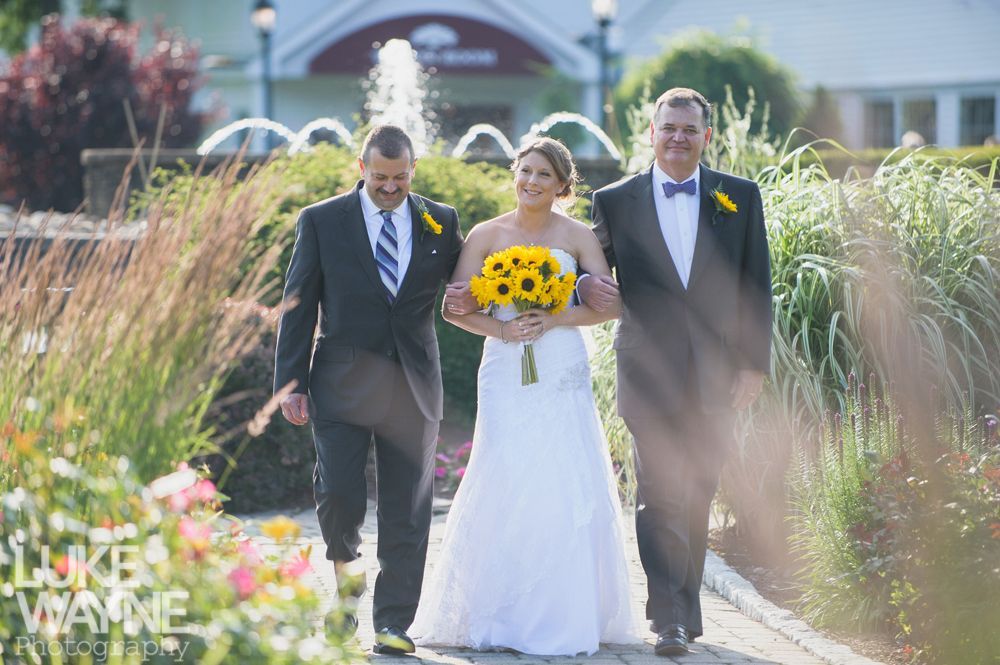 Bride walks down a path with two men, holding a yellow bouquet. Fountain and building in the background.